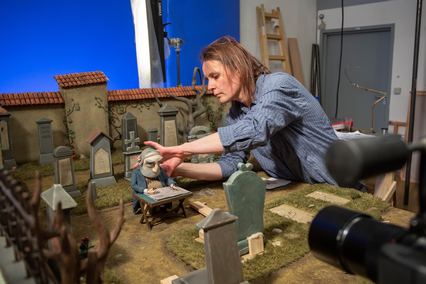 Ivana Bošnjak sen agiba nad scenografijo, v kateri Bogdan sedi za mizico, postavljeno pred grob na pokopališču, in premika lutko.