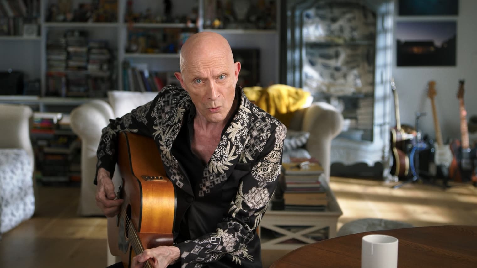 Richard O'Brien, oblečen v glam rock kostum in s kitaro v roki