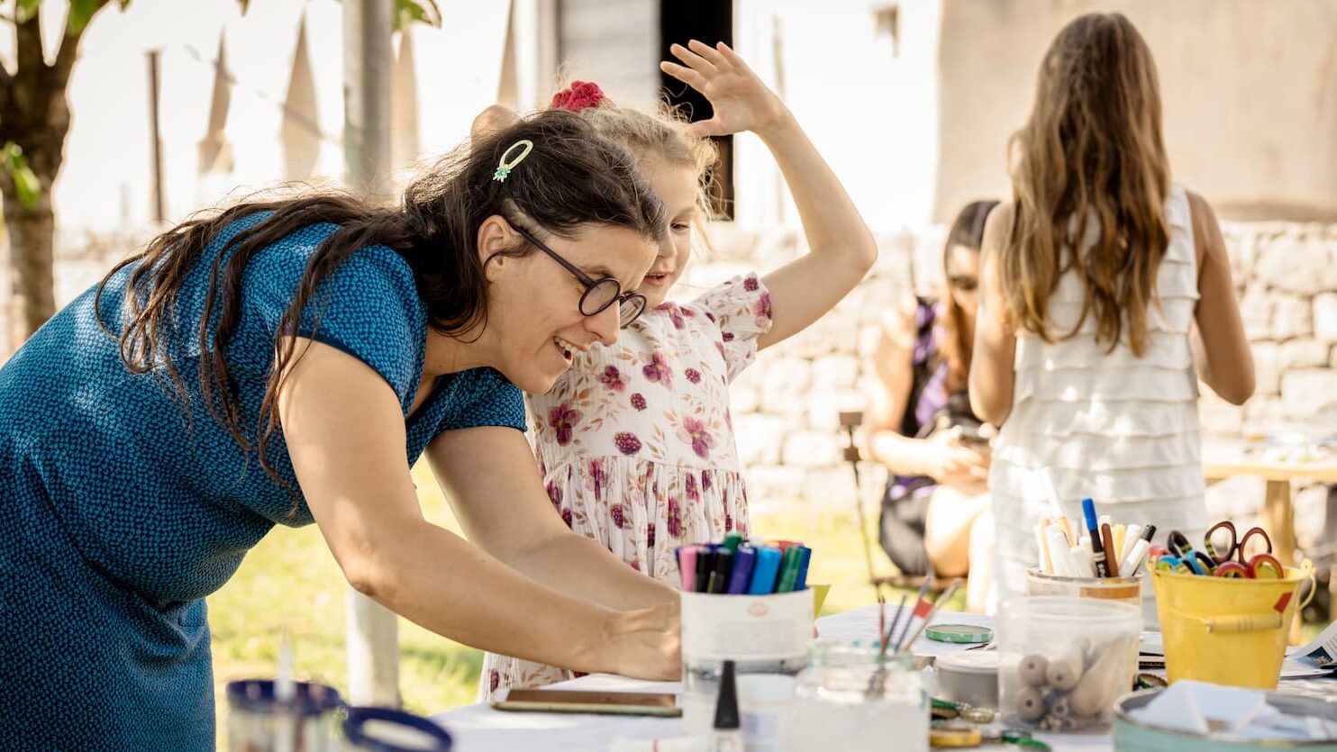 mentorica pomaga deklici ob polni mizi potrebščin za ustvarjanje